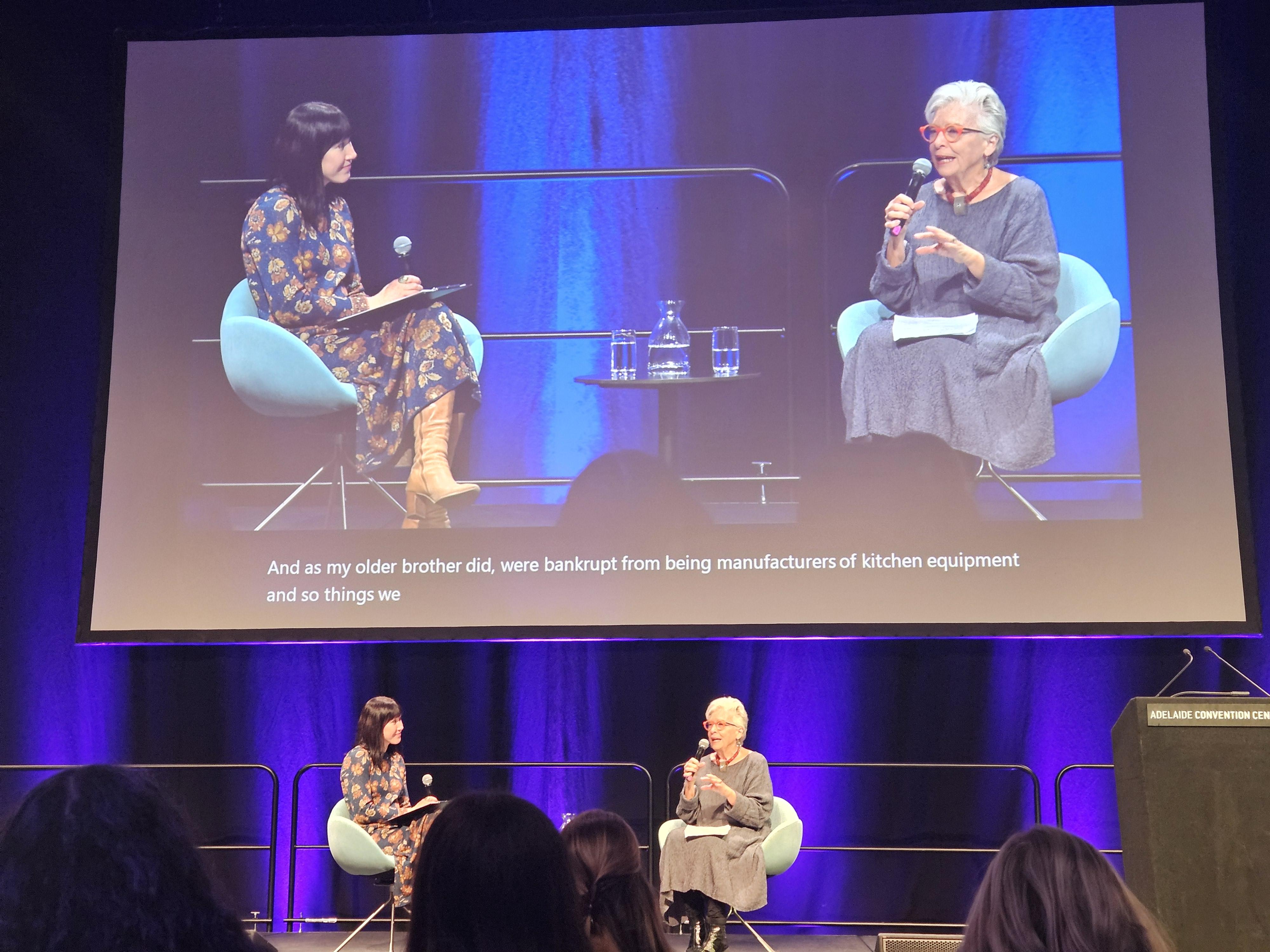 This is a photo of Associate Professor Jade Cartwright and Maggie Beer AO sitting on chairs on stage in conversation. There is a large projector at the back with the subtitles that say 'And, as my older brother did, were bankrupt from being manufacturers of kitchen equipment and so things we...'