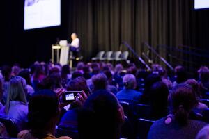 Rows of attendees watching a presenter, taken from behind, one attendee holding up their phone to take a picture of slides