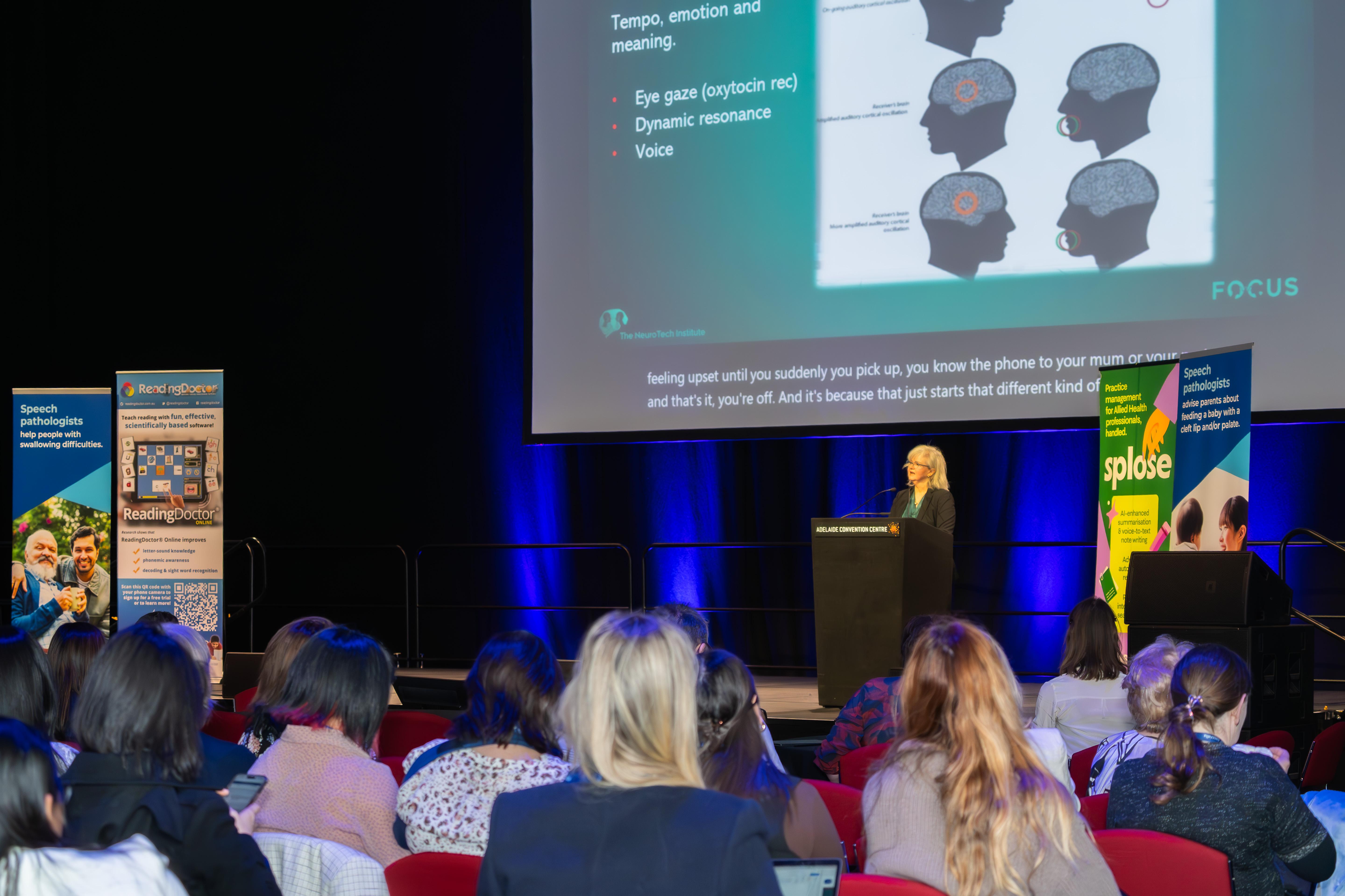 This is a photo of Dr Fiona Kerr standing on stage behind a podium, presenting to the hall of people. Her presentation is being displayed on a large screen at the back. 