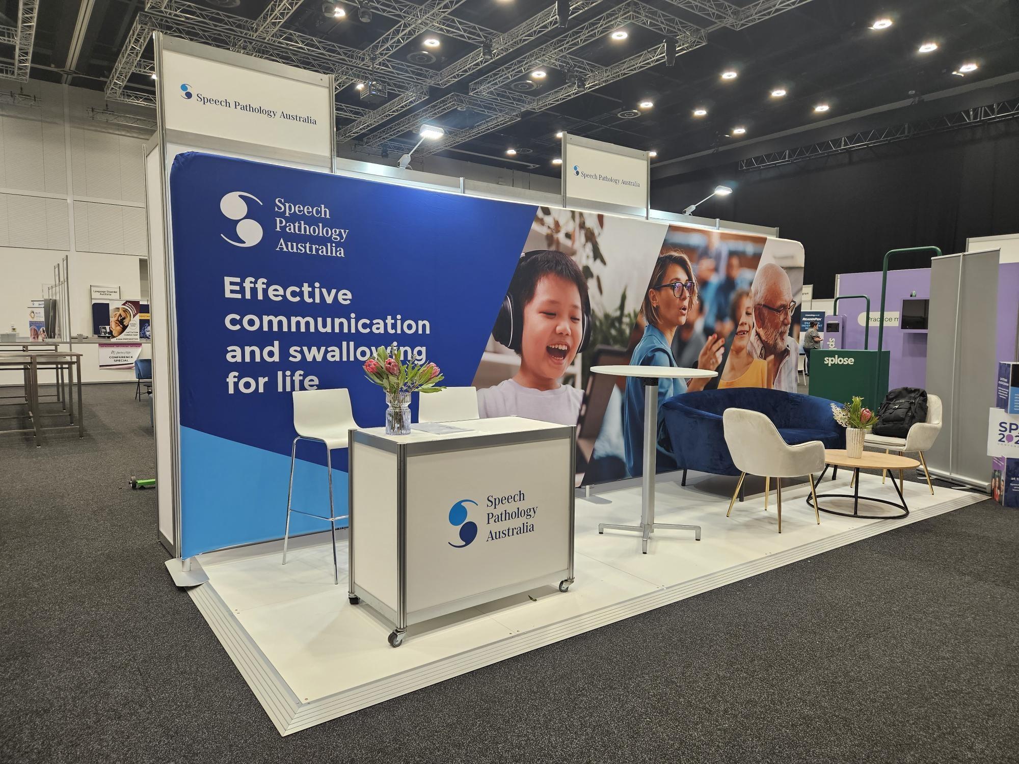 This is a photo of a stall. It shows a large backdrop with various images including a woman giving a speech. There is text that says 'Effective communication and swallowing for life' with the SPA logo above it. In the stall, you can see tables and chairs and SPA staff talking to conference goers. In the foreground there are conference goers taking food from the catering table. 