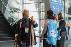 An attendee speaking to two student volunteers wearing blue bibs, one holding a large lollipop sign.