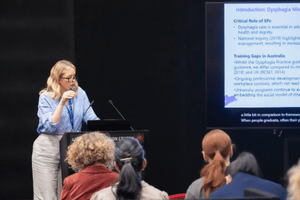 A presenter at a lectern presenting to rows of attendees, with a TV behind displaying their presentation with subtitles below. 